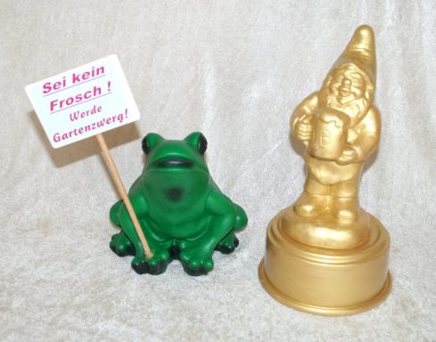 Grüner Frosch mit Schild und goldener Gartenzwerg auf einem Tisch.